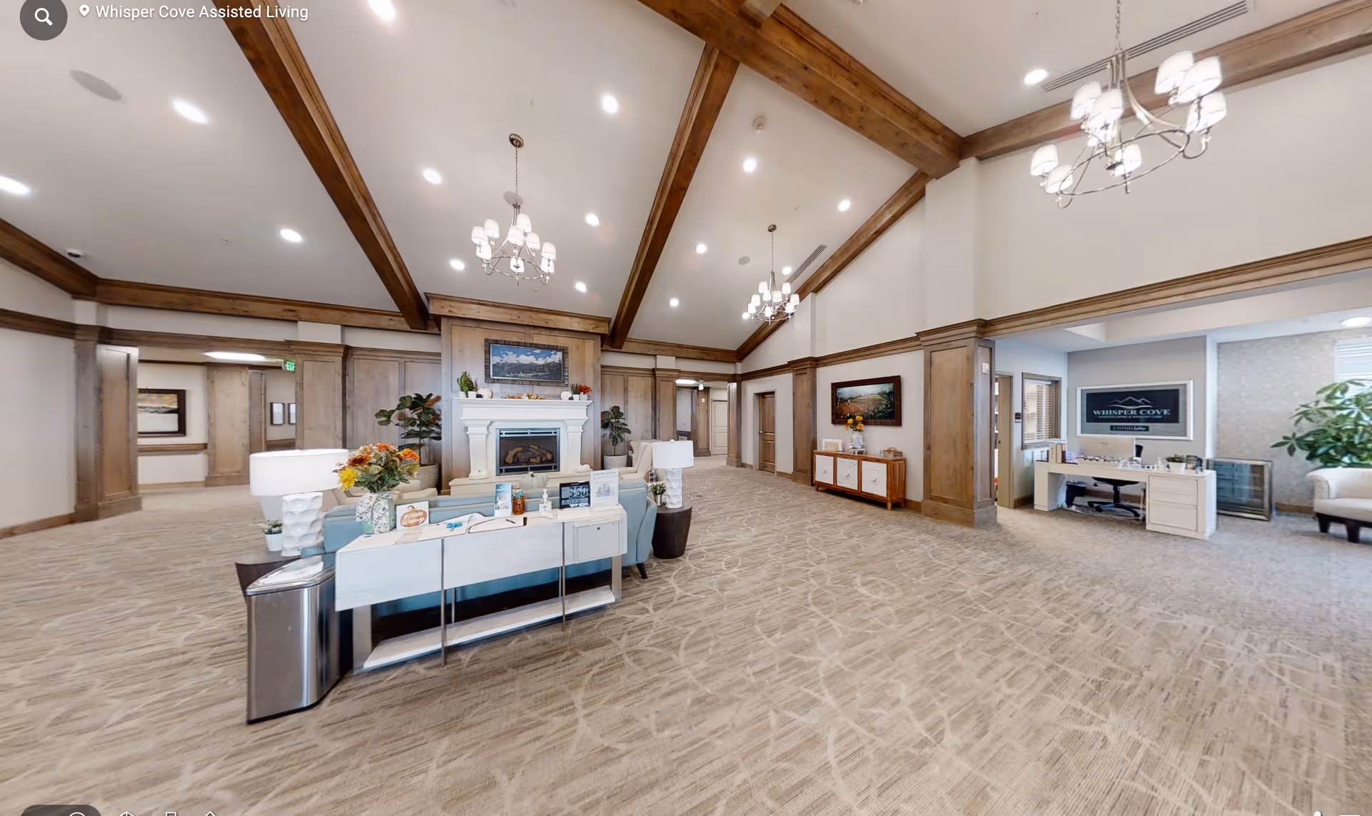 Spacious assisted living lobby with vaulted beamed ceiling, chandeliers, seating area and a central fireplace.