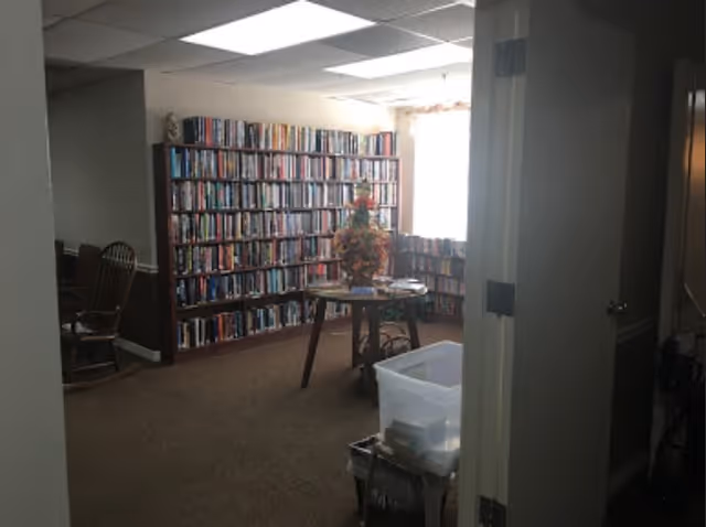 Interior view of a room with bookshelves filled with books along the walls, a round table with a floral arrangement in the center, and a rocking chair to the left. The room is carpeted and lit by natural light from a window with sheer curtains.
