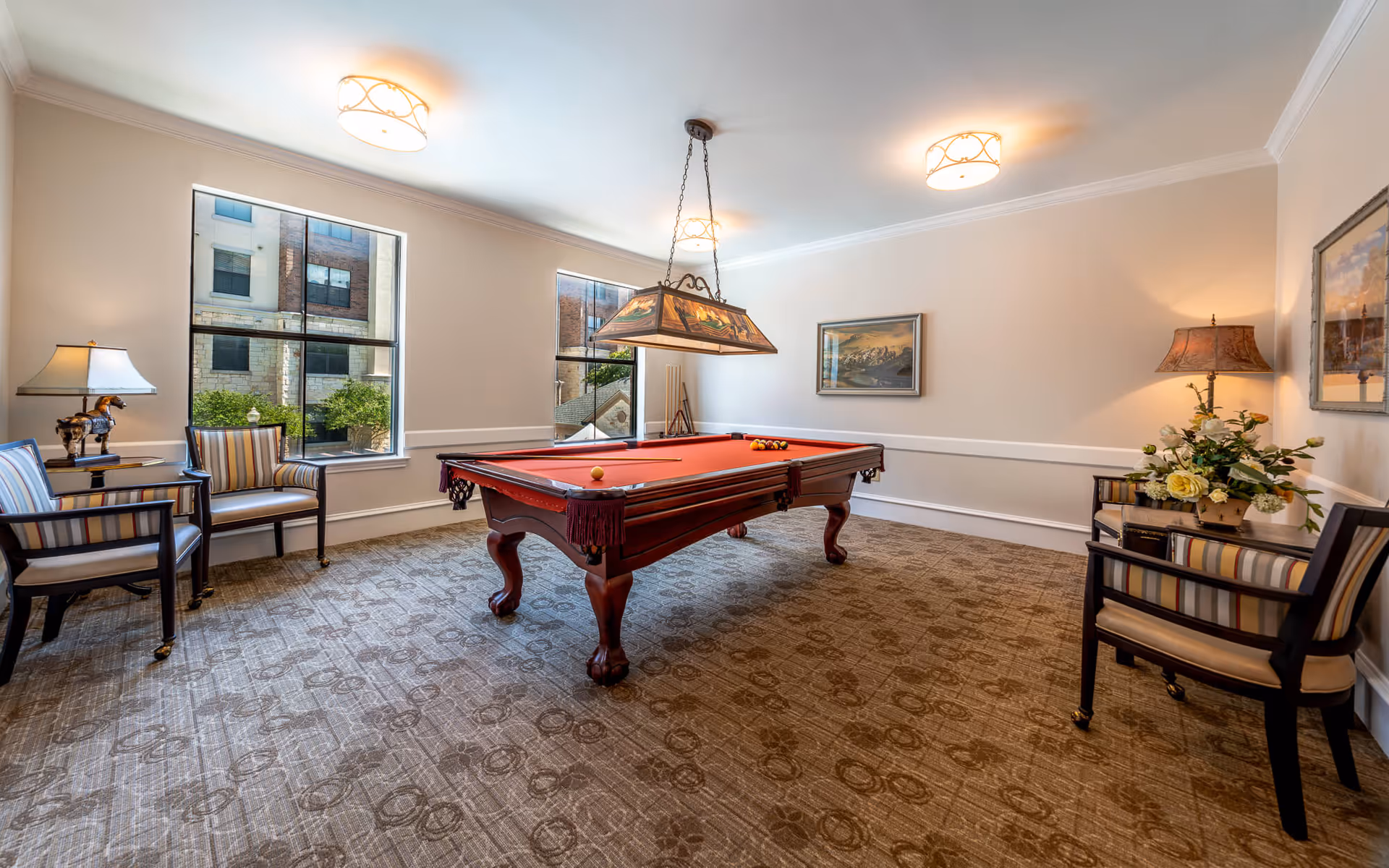 A well-lit game room with a red felt pool table in the center, surrounded by four striped armchairs. The room has beige walls, two large windows showing an exterior building, framed paintings on the walls, and decorative lamps providing warm lighting. The carpet has a patterned design.