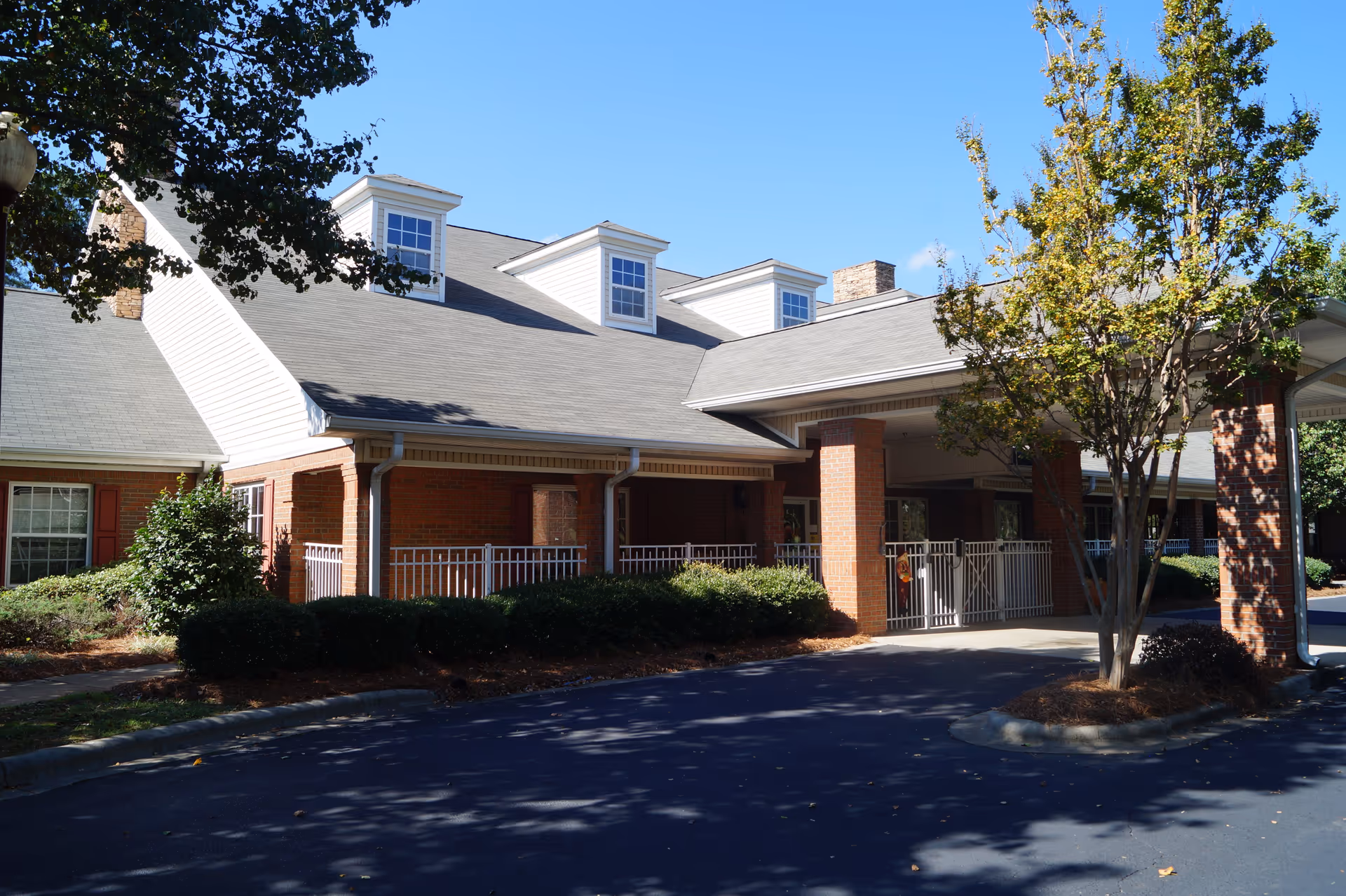 Exterior view of a senior living facility building with brick walls, white trim, and a covered entrance supported by brick columns. There are bushes and a tree in front of the building under a clear blue sky.