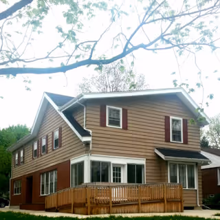 Two-story tan-sided house with brown shutters and a wooden front ramp/deck under tree branches.