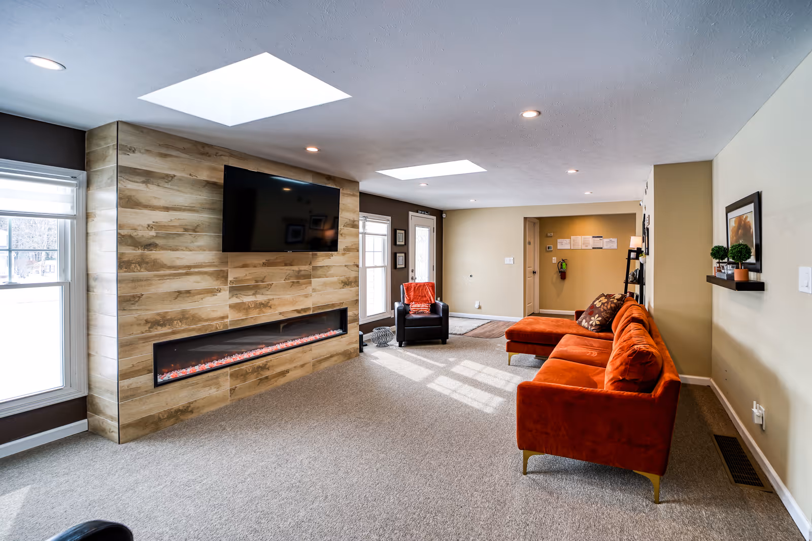 A bright living room with beige walls and carpeted floor featuring a modern wood-paneled wall with a mounted flat-screen TV and a long electric fireplace below it. The room has two skylights, a large window, an orange sectional sofa with decorative pillows, a black armchair with an orange throw, and a small shelf with potted plants and framed artwork on the wall.