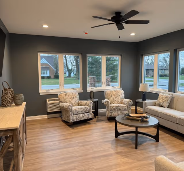 Cozy common living room with two patterned armchairs, a sofa, round coffee table, side tables, ceiling fan, and multiple windows.