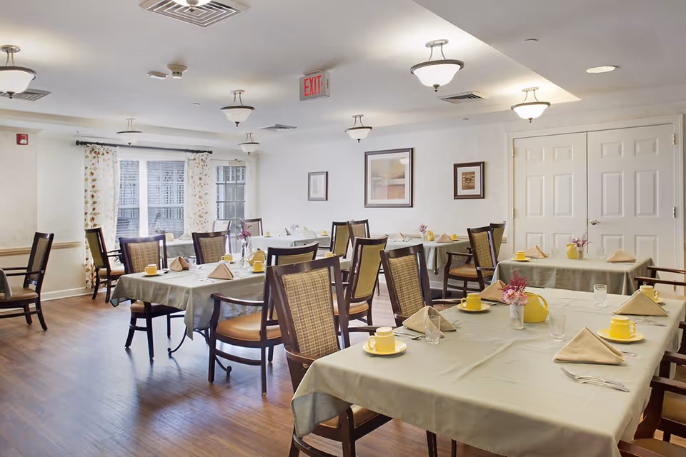 A bright dining room with several tables and chairs set with yellow cups, folded napkins, and small centerpieces.