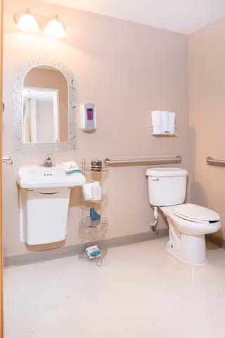 A clean bathroom with a white wall-mounted sink and a decorative mirror above it. To the right of the sink is a white toilet with a grab bar on the wall behind it. There is a metal rack holding toiletries and a soap dispenser mounted on the wall above the sink. The walls are painted beige and the floor is light-colored.