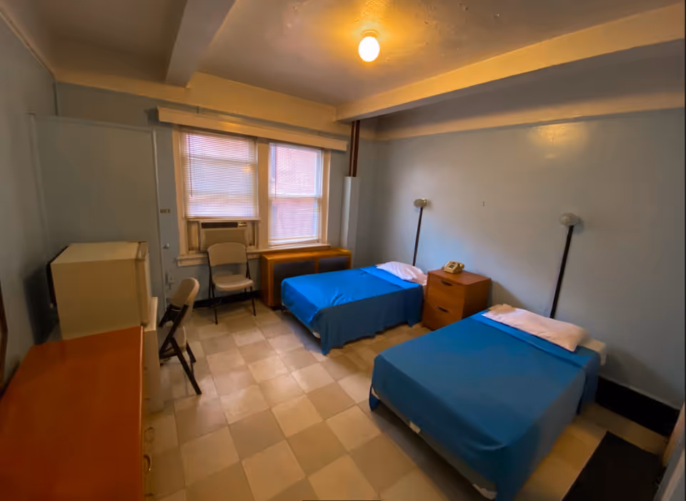 A simple room with two single beds covered in blue bedspreads, each with a white pillow. Between the beds is a small wooden nightstand with a telephone on it. The room has light blue walls, a tiled floor, two windows with blinds, an air conditioning unit, two chairs, a small refrigerator, and a wooden dresser.