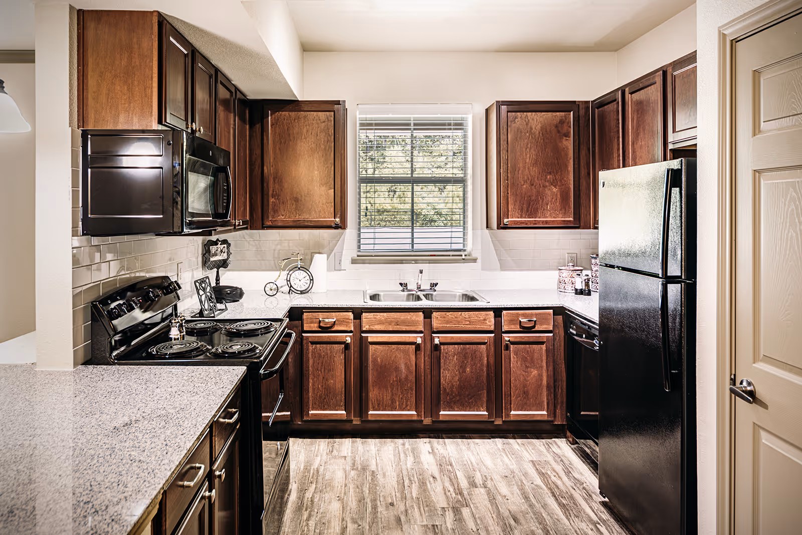 A modern kitchen with dark wooden cabinets, a black refrigerator, black stove, black microwave, and a double sink under a window with blinds. The countertops are light-colored granite, and the floor has a wood-like finish.