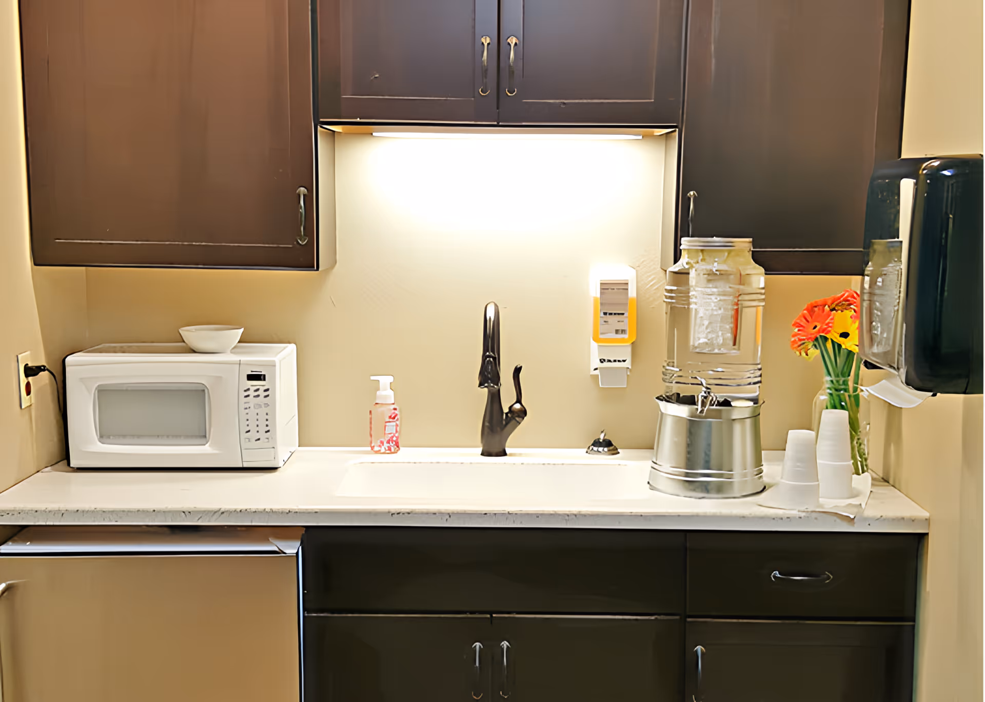 A kitchenette area with dark brown cabinets, a white countertop, a white microwave, a black faucet over a sink, a soap dispenser, a water dispenser with cups, and a vase with orange and yellow flowers.