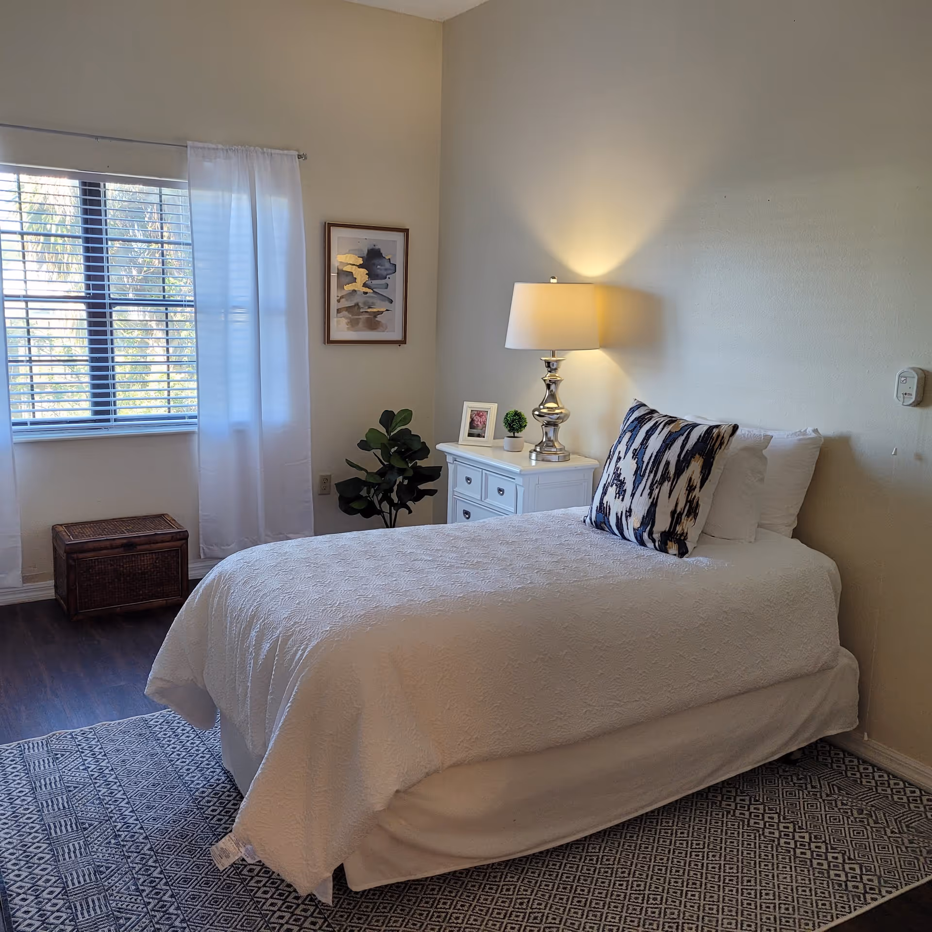 A neatly made single bed with white bedding and a decorative pillow in a bright bedroom. Next to the bed is a white nightstand with a silver lamp, a small plant, and a framed photo. A window with white curtains and blinds lets in natural light. There is a piece of abstract art on the wall and a wicker basket on the floor near the window. The floor is dark wood with a patterned rug under the bed.
