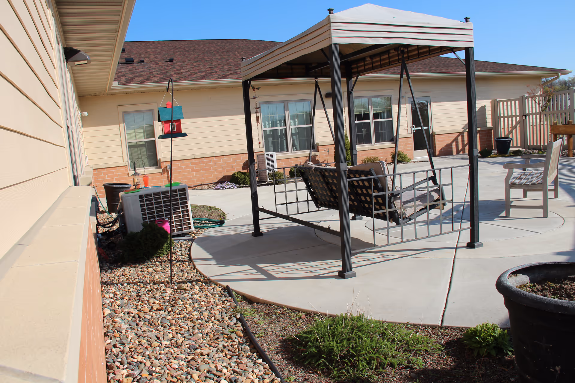 Outdoor patio area at Villages of Lonsdale by Three Links with a metal swing under a canopy, chairs, a bird feeder, and landscaping including rocks and plants. The building exterior is visible with beige siding and brick accents.