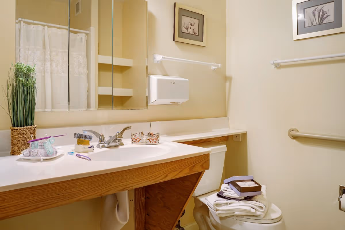A small accessible bathroom with a sink and vanity, mirrored medicine cabinet, toilet topped with folded towels and wall-mounted grab bars.