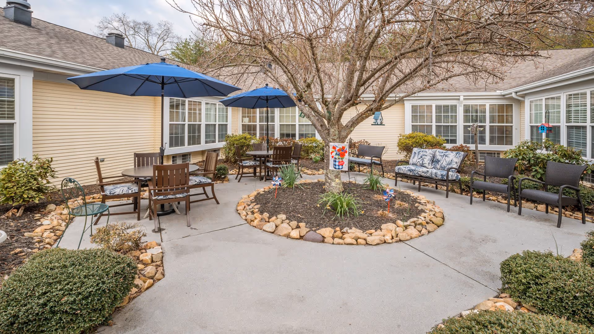 Outdoor courtyard area with a circular garden bed featuring a tree in the center, surrounded by patio seating including chairs, benches, and tables with blue umbrellas. The courtyard is bordered by a beige building with multiple windows and some shrubs and plants around the edges.