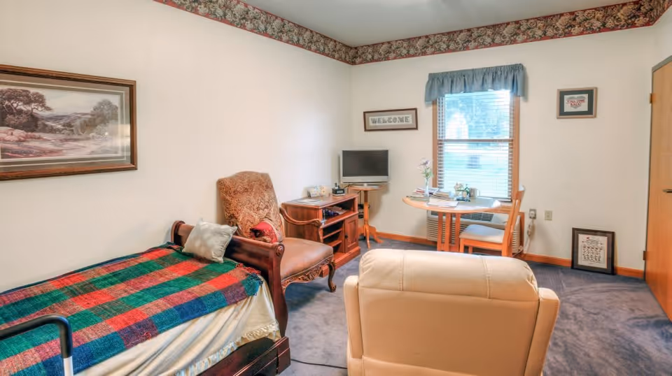 A cozy senior living bedroom with a single bed covered by a red, green, and blue plaid blanket. There is a cushioned armchair next to the bed, a small wooden TV stand with a flat-screen TV, and a round wooden table with two chairs near a window with blue valance curtains. The walls are white with floral wallpaper border near the ceiling, and there are framed pictures and a 'WELCOME' sign on the walls.