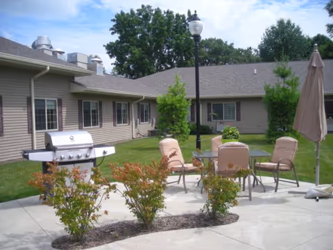Patio courtyard with an outdoor dining table and chairs, a grill, umbrella, and shrubs in front of a single-story senior living building.