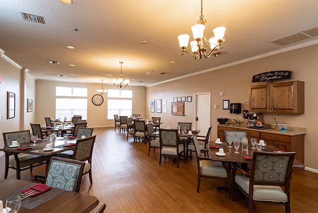 A well-lit dining room with multiple wooden tables and chairs arranged neatly. Each table is set with placemats, napkins, glasses, and cups. The room has wooden flooring, beige walls, and several windows letting in natural light. There are chandeliers hanging from the ceiling and framed pictures on the walls. A small counter area with cabinets and a coffee machine is visible on the right side.