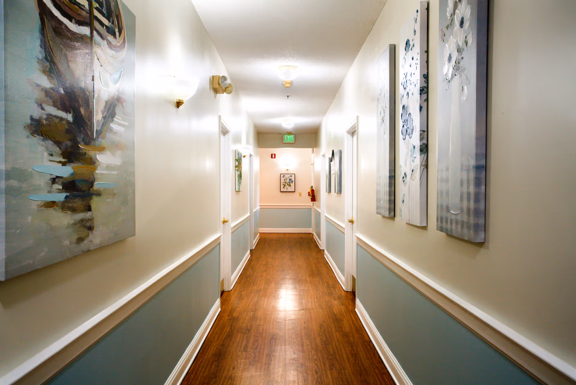 A well-lit hallway in an assisted living facility with wooden flooring, light-colored walls with blue wainscoting, and several doors on both sides. The walls are decorated with framed artwork and floral paintings. An exit sign and fire extinguisher are visible at the end of the hallway.