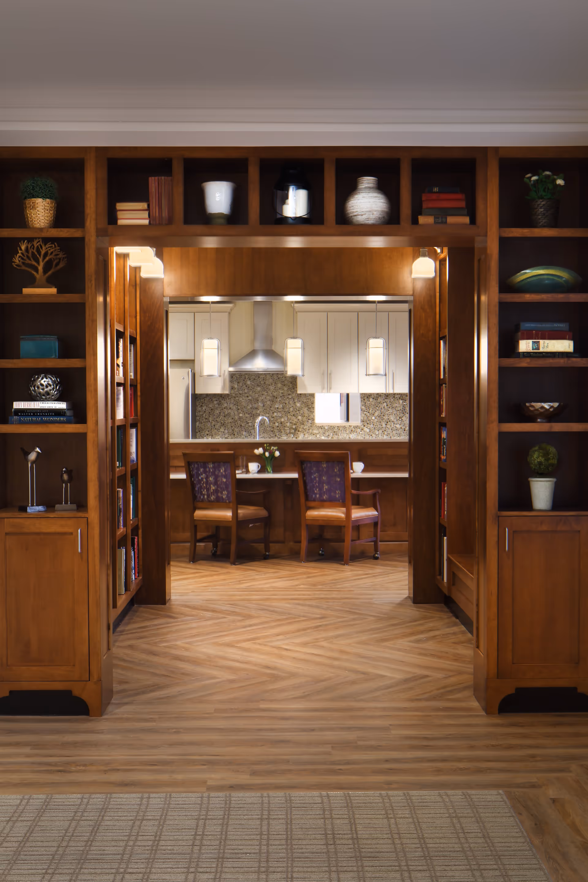 View through a wooden bookshelf-lined hallway into a kitchen area with white cabinets, a granite backsplash, and a stainless steel range hood. Two wooden chairs with purple cushions are positioned at a counter with a vase of flowers and cups. The floor is wood with a herringbone pattern.