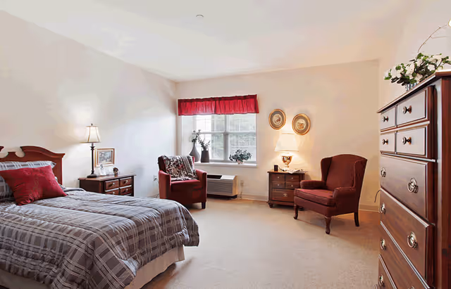 A cozy bedroom with a bed covered in a plaid comforter and red pillows, two red upholstered armchairs near a window with a red valance, two wooden nightstands with lamps and decorative items, and a tall wooden dresser with plants on top.