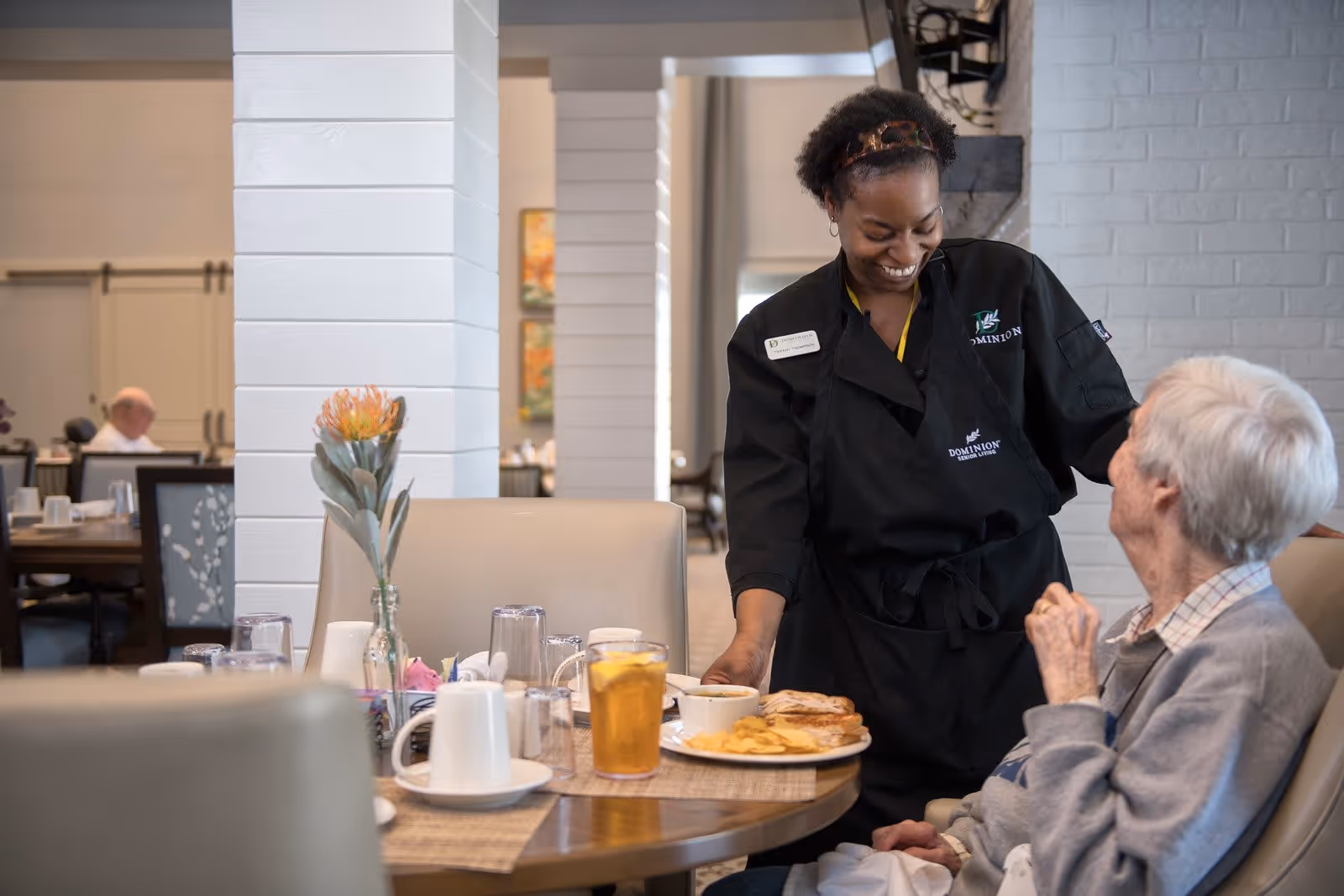 A smiling staff member in a black Dominion Senior Living uniform serves food to an elderly woman seated at a dining table in a senior living facility. The table has plates with food, glasses, and a flower in a vase. Other residents are visible in the background in a dining area.