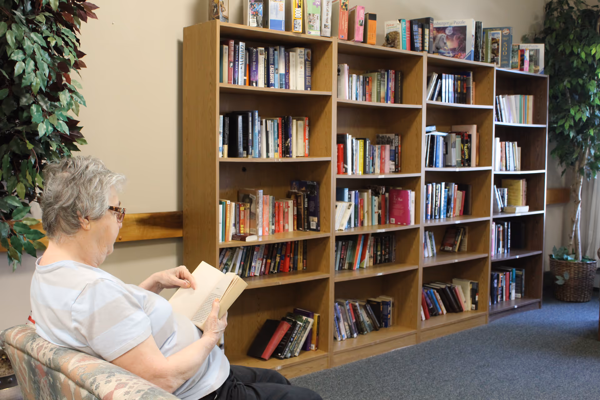 An elderly woman with short gray hair and glasses is sitting on a patterned couch reading a book in a room with multiple wooden bookshelves filled with books. There are two large potted plants on either side of the bookshelves, and the room has a carpeted floor.