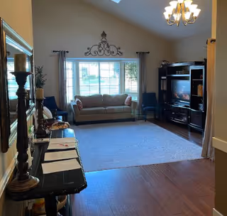 Sunlit living room with a sofa beneath a large window, chairs, a TV console, and a rug on hardwood floors.