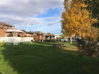 Outdoor area with green grass, a wooden picnic table, two gazebos, and a white fence. There are buildings in the background and a tree with yellow autumn leaves on the right side under a partly cloudy sky.