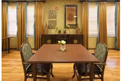 Dining room with a wooden table and four upholstered chairs, a sideboard against a wallpapered wall and tall windows with yellow curtains.
