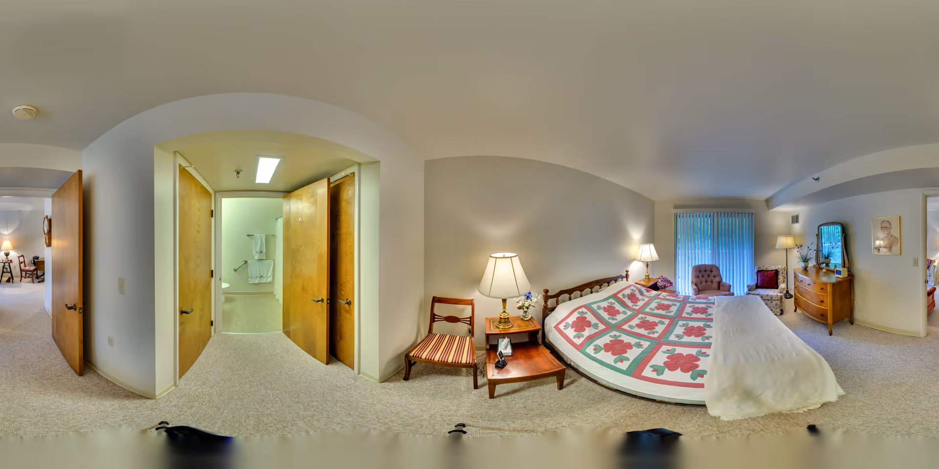 A spacious bedroom in The Lodge Retirement Community featuring a large bed with a floral quilt, two bedside tables with lamps, a wooden chair with a striped cushion, a dresser with a mirror, and a sitting area with two armchairs near a window with vertical blinds. The room has beige carpet and walls, and an open doorway leads to a bathroom with towels hanging on the wall.