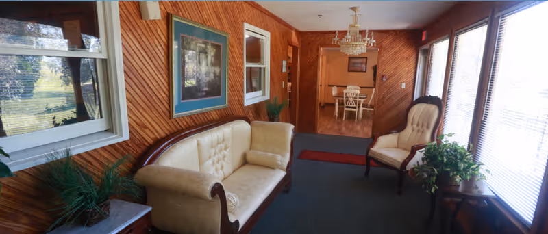 Wood-paneled common sitting area with an upholstered sofa and chairs beside large windows and a view into a dining area.
