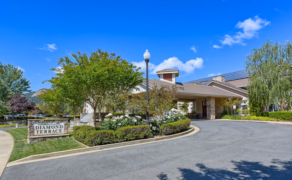 Exterior view of Diamond Terrace retirement living facility with a driveway, landscaped greenery, trees, and a clear blue sky.