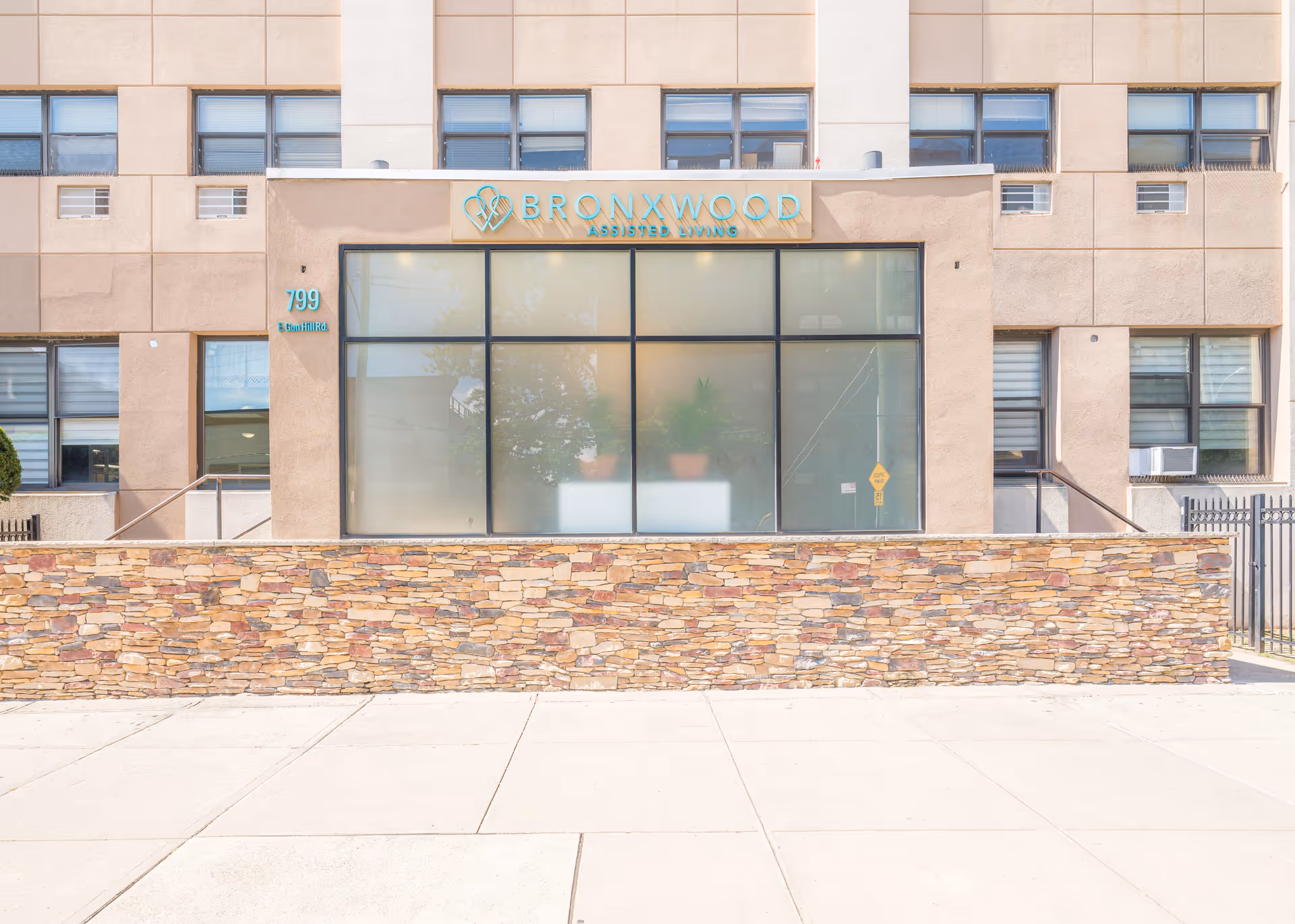 Front exterior view of Bronxwood Assisted Living facility showing a beige building with large windows and a stone wall in front. The facility name and logo are displayed above the windows, along with the address number 799 E. Gun Hill Rd.