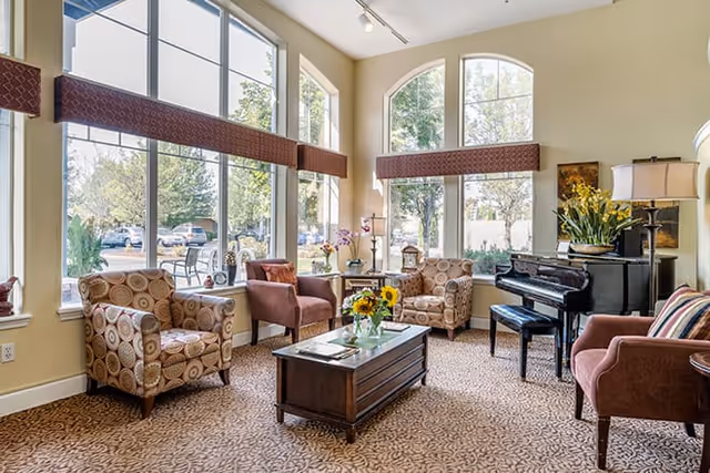 A bright and inviting living room with large arched windows letting in natural light. The room features patterned armchairs, a brown leather chair, a wooden coffee table with a vase of sunflowers, a black piano with a matching bench, and decorative plants and lamps. The carpet has a subtle pattern, and the walls are painted a soft beige.