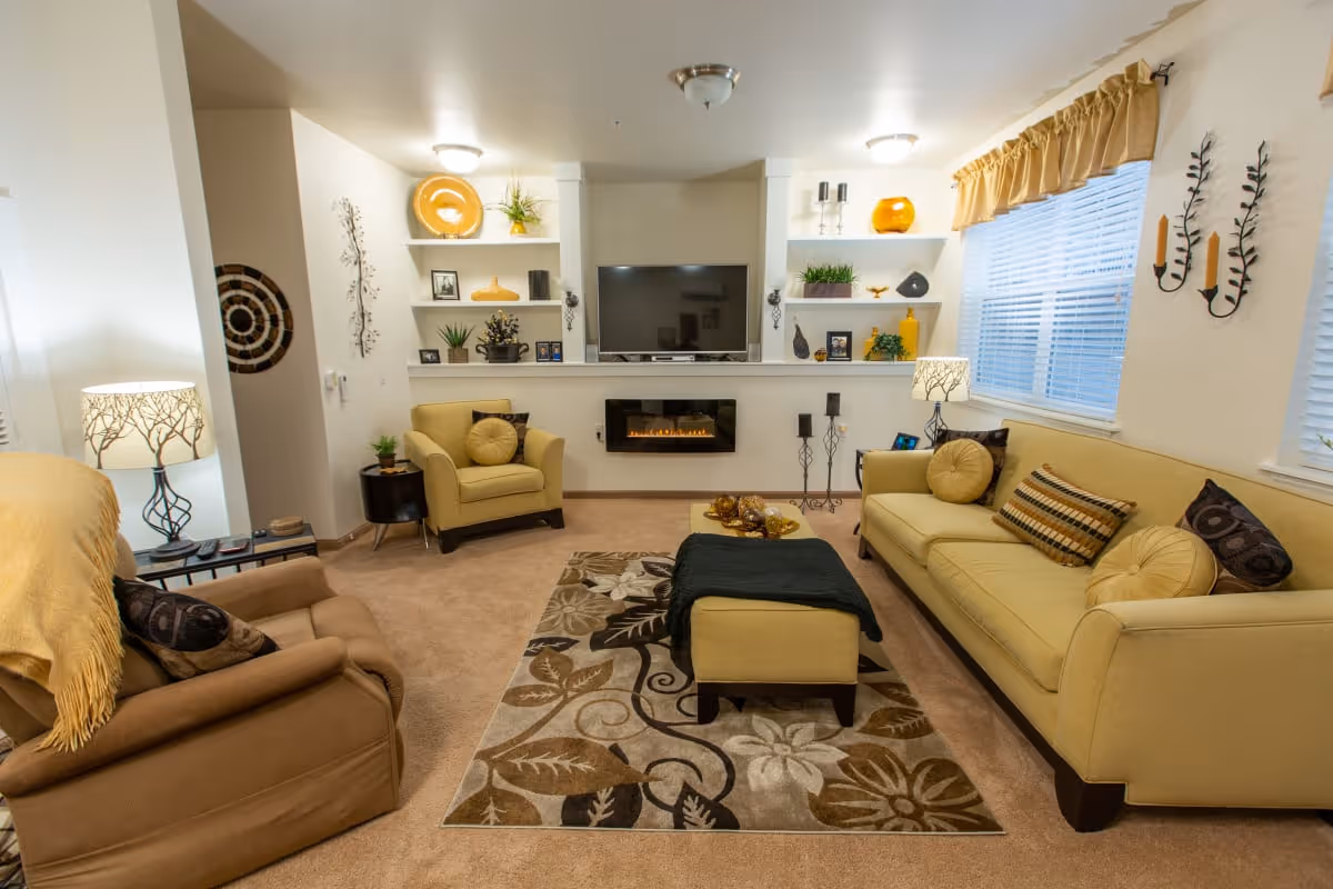 A cozy living room with beige carpet and light yellow walls. The room features a yellow sofa with multiple cushions, a matching armchair, and a brown recliner chair with a yellow throw blanket. A patterned area rug lies in the center with a small ottoman covered by a dark throw. The wall opposite the sofa has built-in shelves decorated with plants, vases, and framed photos, with a flat-screen TV mounted above a modern electric fireplace. Two table lamps with tree-patterned shades provide warm lighting, and two windows with blinds and yellow valances let in natural light. Wall-mounted candle holders and decorative items add to the inviting atmosphere.