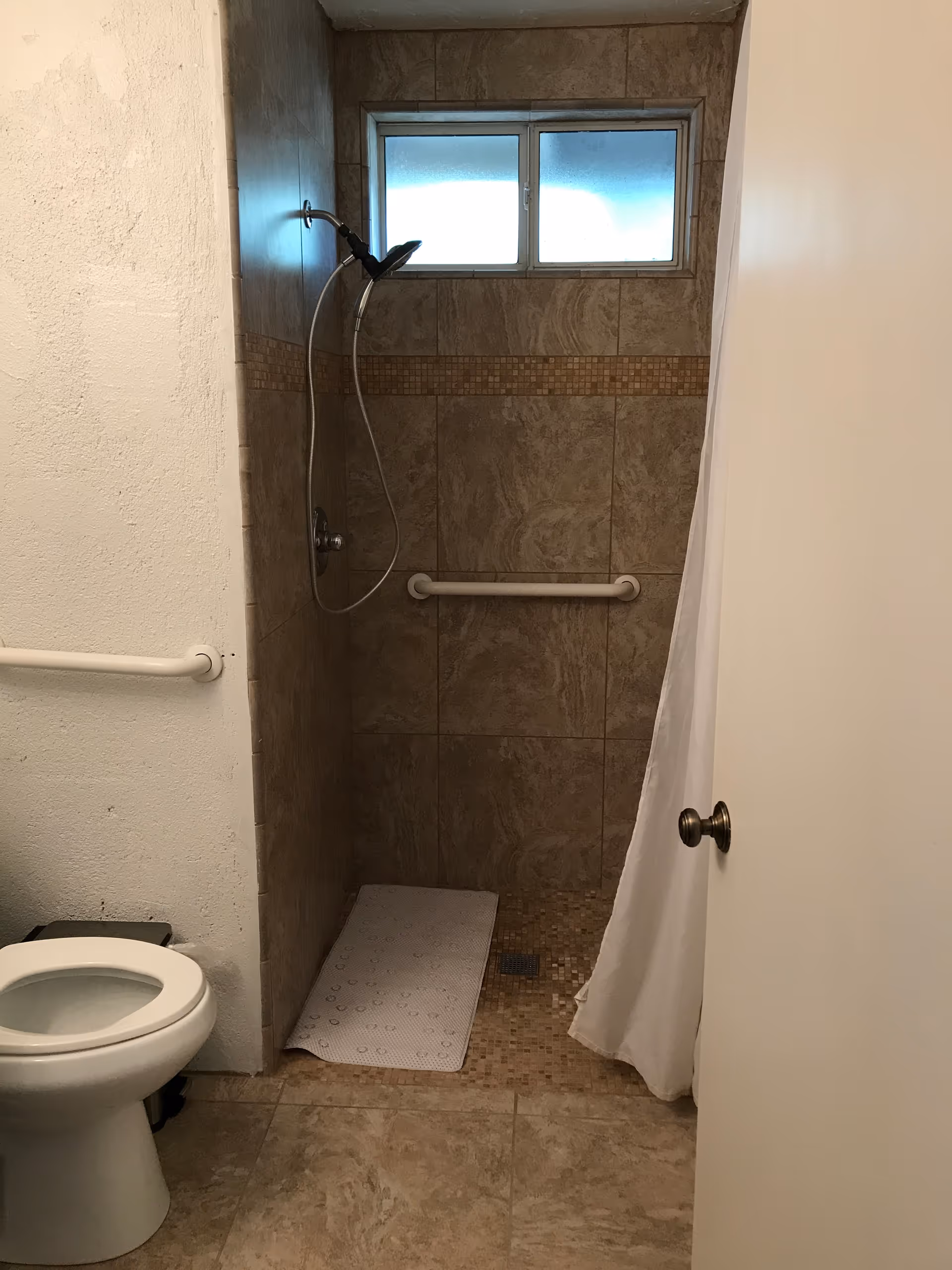 A bathroom with a tiled walk-in shower featuring a handheld showerhead, a grab bar on the wall, and a white shower curtain partially open. There is a small frosted window above the shower. To the left, a white toilet is visible with a grab bar mounted on the wall beside it. The floor is tiled in a beige color.