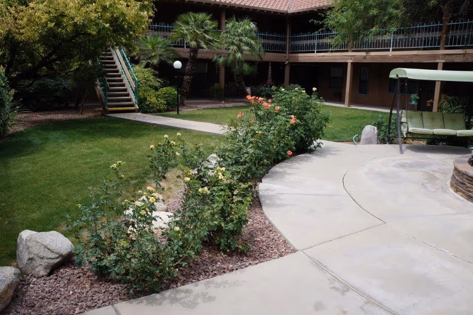 Courtyard with paved walkways, rose bushes, lawn, a green cushioned swing, and a two-story building with exterior balconies.