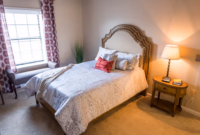 Cozy bedroom with a made bed featuring decorative pillows, a wooden headboard, a nightstand with a lamp, and a window with patterned curtains.