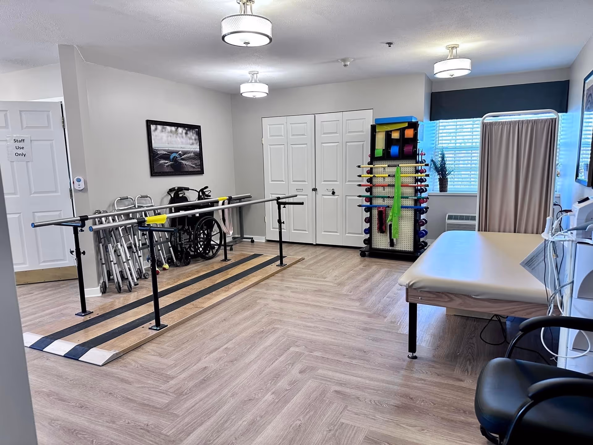 A bright therapy room with parallel walking bars, walkers and a wheelchair, an exercise equipment rack, and a treatment table.
