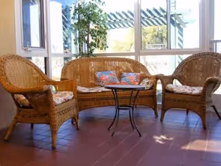 A sunlit seating area with wicker furniture including a loveseat and two armchairs, all with floral cushions, arranged around a small round glass-top table. The area has large windows with a view of greenery outside.