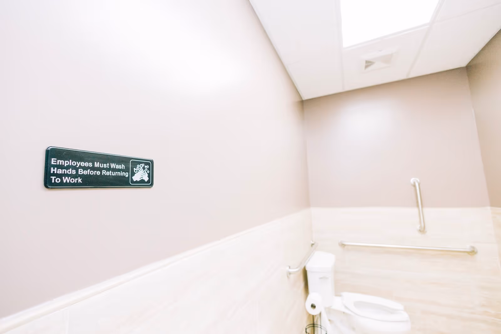 A clean, accessible bathroom with beige walls and tiled lower walls. There is a white toilet with grab bars on the walls for support. A sign on the wall reads 'Employees Must Wash Hands Before Returning To Work.'