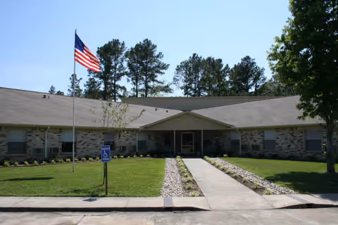 Single-story brick building with a sloped roof, an American flag on a flagpole, a sidewalk leading to the entrance, and a well-maintained lawn with trees and shrubs.
