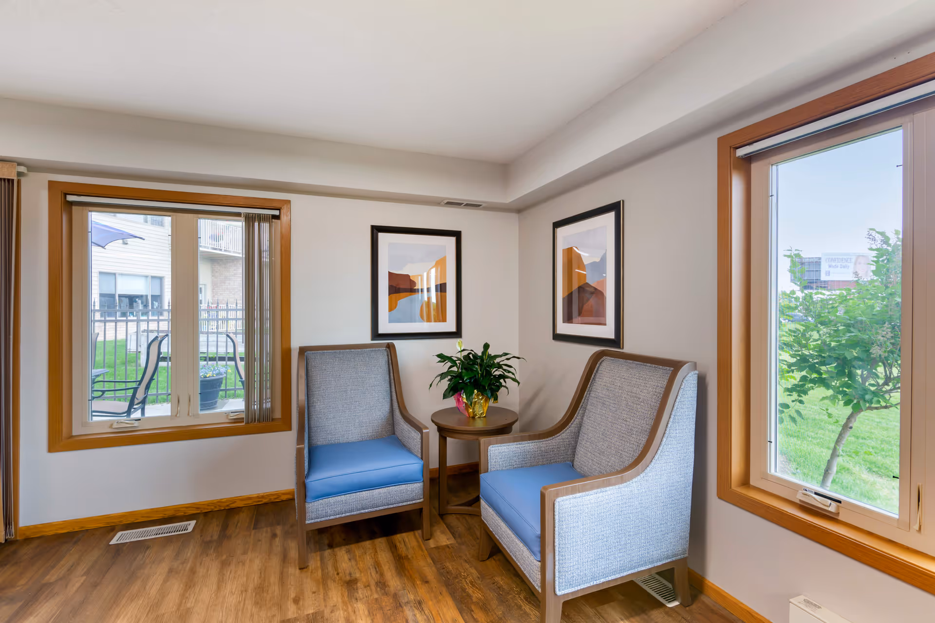 A cozy sitting area in a senior living facility with two cushioned armchairs featuring blue seats and wooden frames, a small round wooden table with a potted plant in the center, two framed abstract art pieces on the wall, and two large windows showing an outdoor view with greenery and patio furniture.