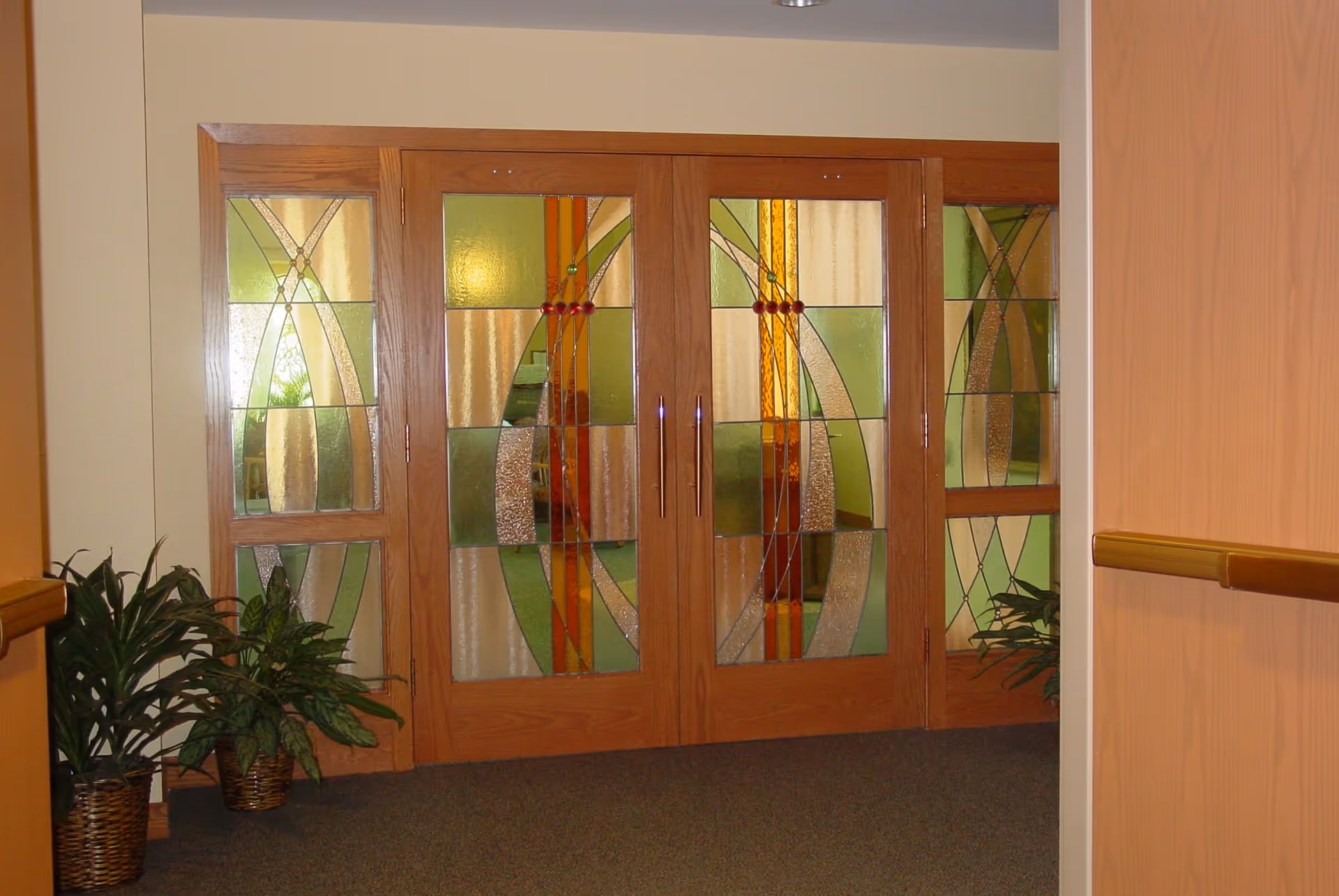 Double wooden doors with decorative stained glass panels featuring green, yellow, and orange geometric patterns. Two potted plants are placed on either side of the doors in a carpeted hallway with beige walls and wooden handrails.