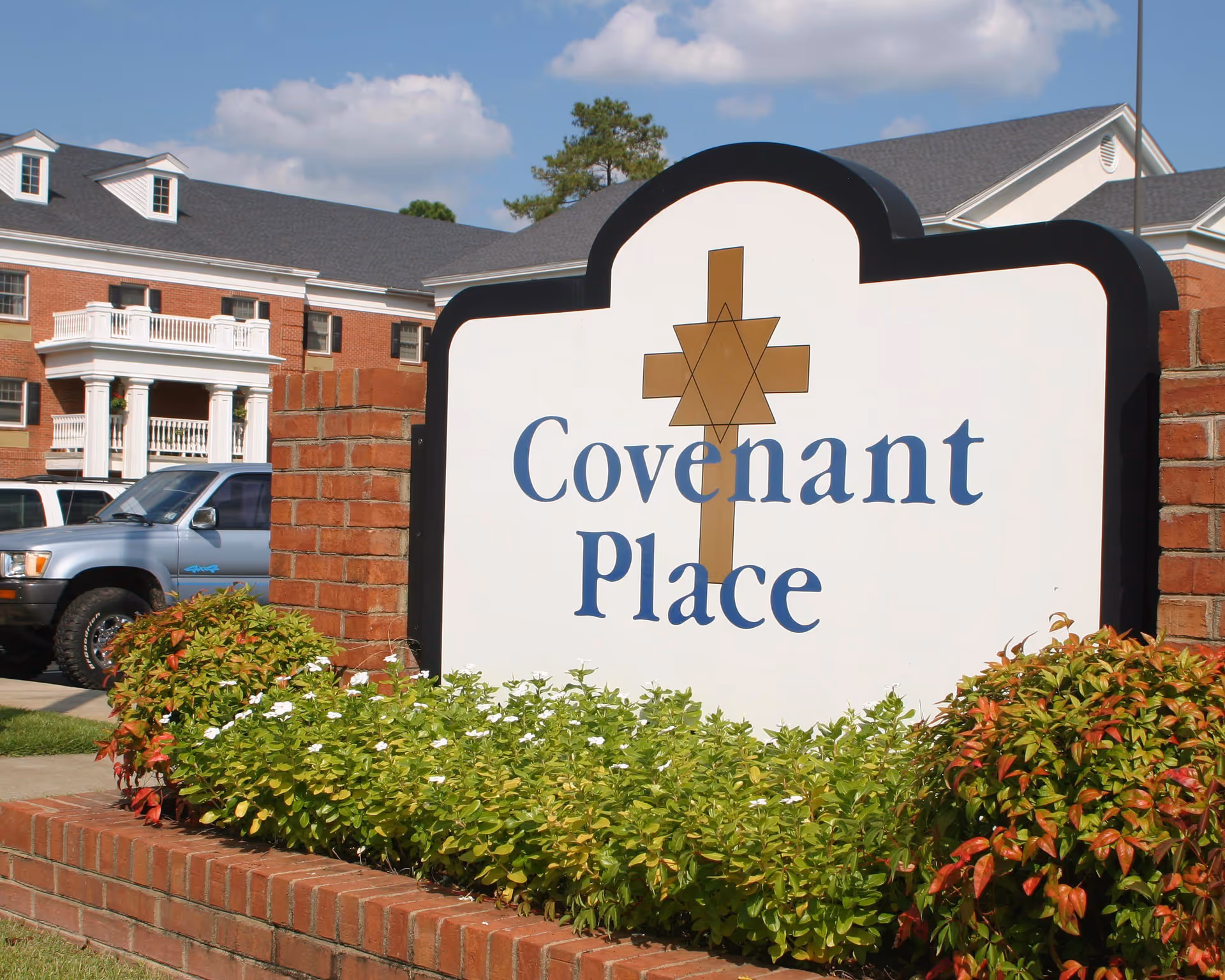 Outdoor view of a senior living facility named Covenant Place with a large sign in the foreground surrounded by green shrubs and a brick base. In the background, there is a red brick building with white balconies and a parked vehicle.