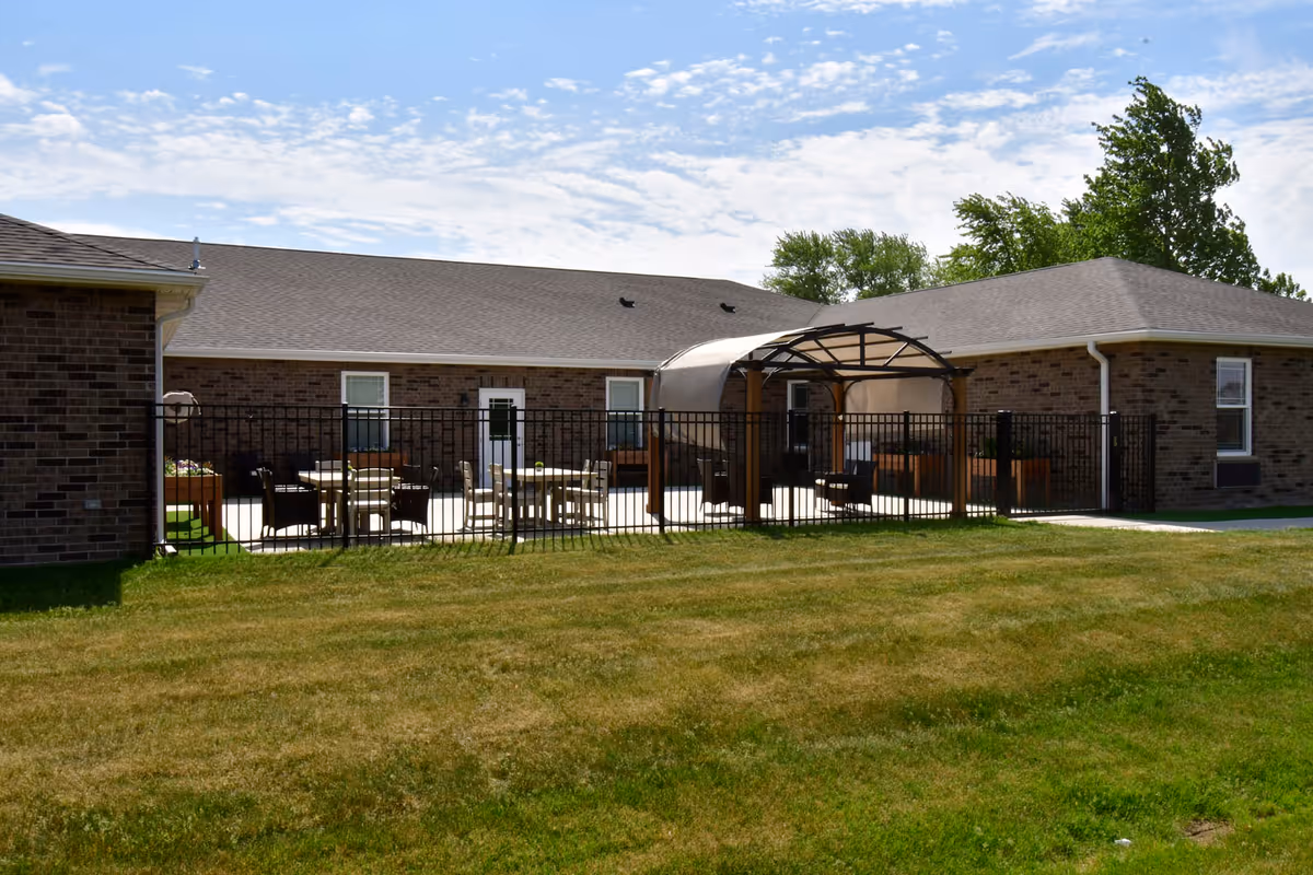 Exterior view of a single-story brick senior living building with a fenced patio, outdoor seating, a canopy, and a grassy lawn.