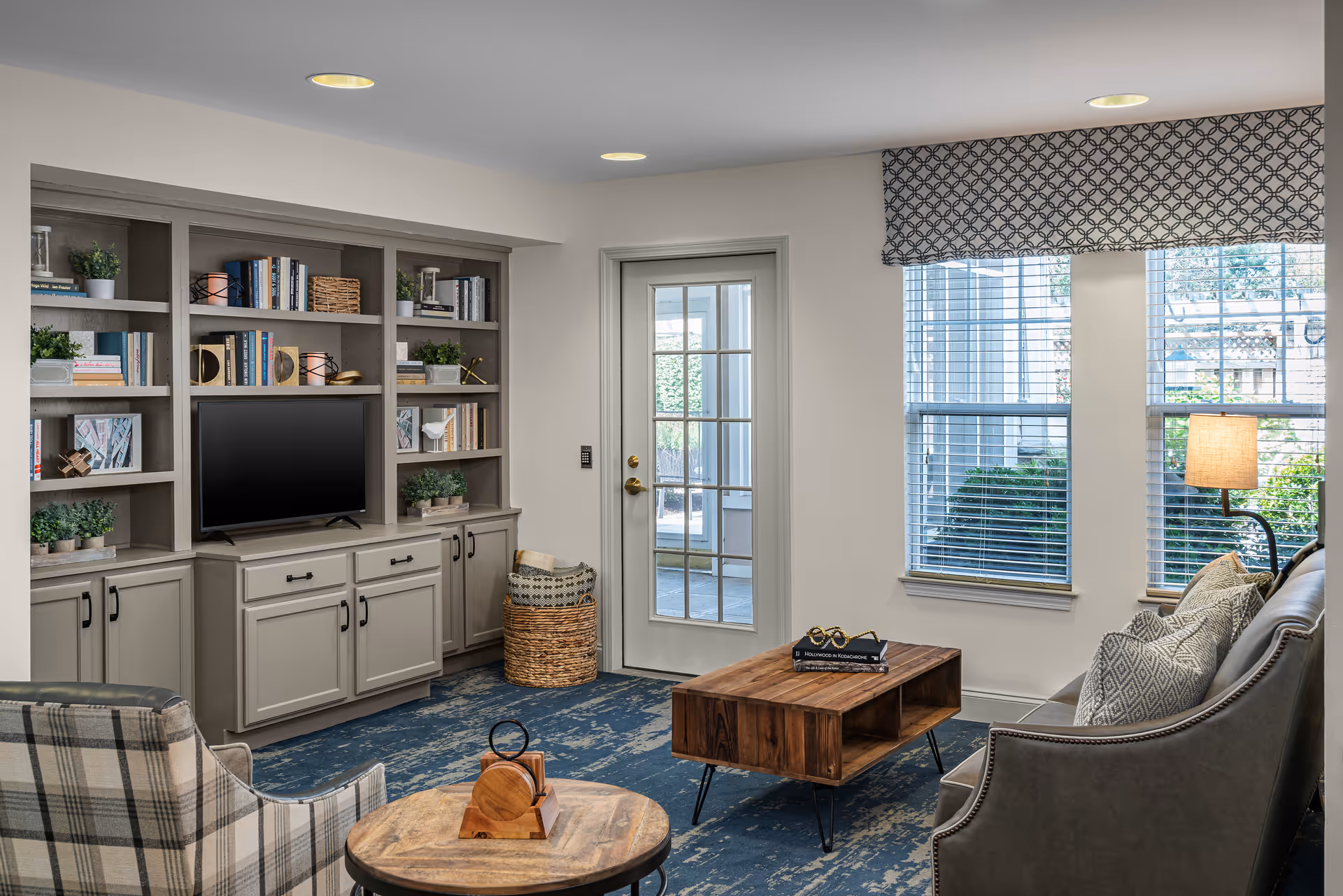 Well-lit living room with built-in bookshelves and a TV, sofa and armchair, wooden coffee table, and large windows.