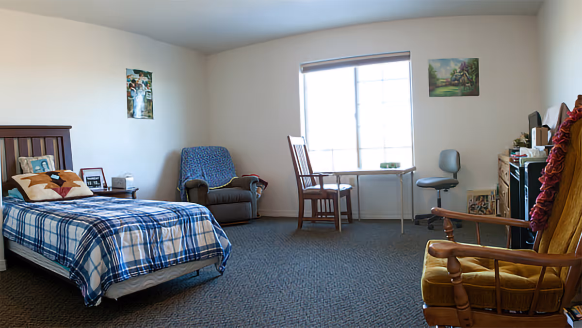 Furnished bedroom with a single bed, armchairs, a small desk by a large window, and carpeted floor.