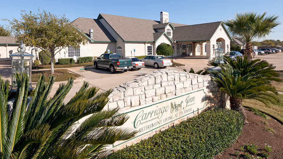 Exterior view of Carriage Inn Lake Jackson retirement living facility showing a stone sign with the facility name, a parking lot with several cars, palm trees, and a single-story building with a pitched roof in the background.