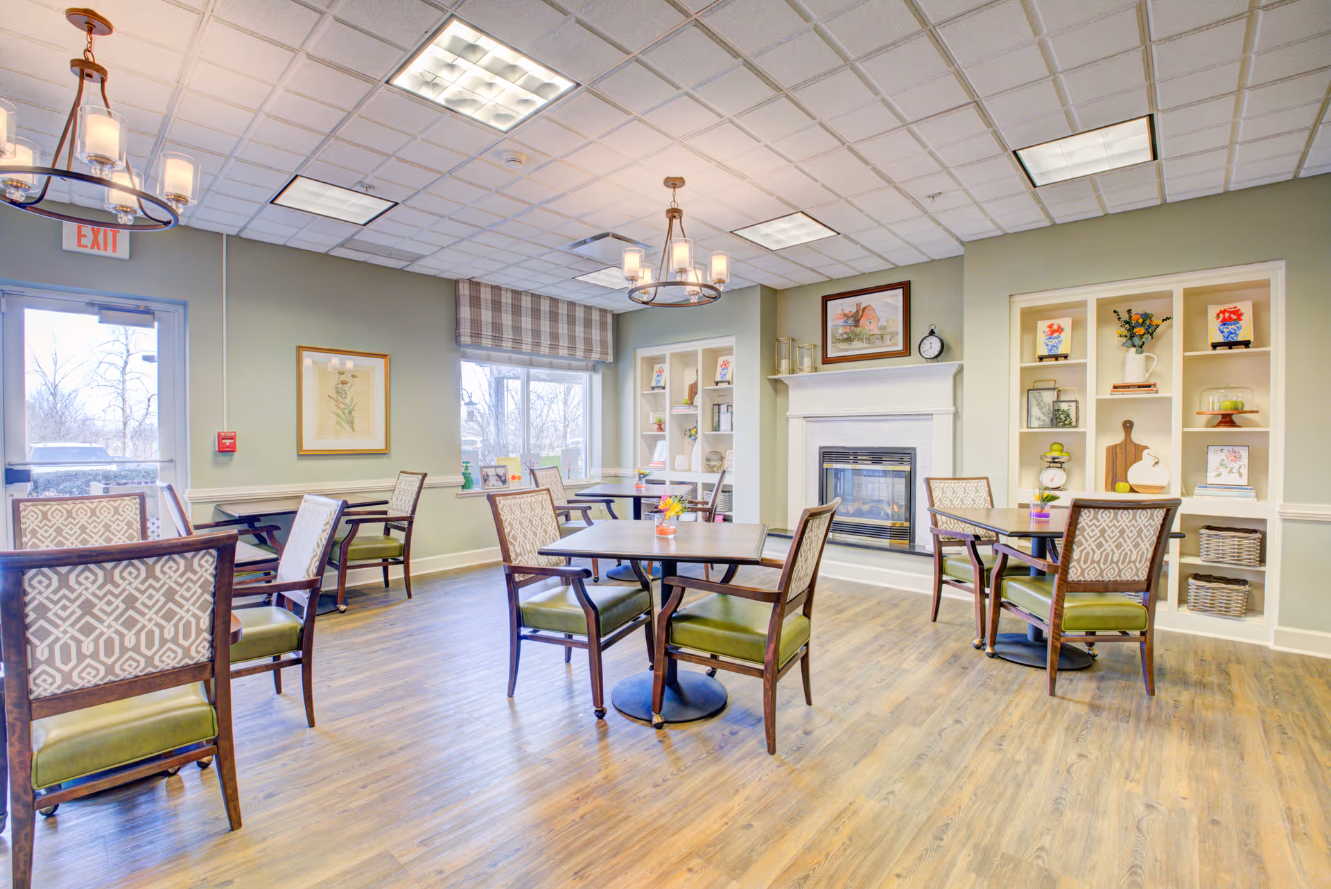 A bright and spacious common area with wooden flooring, several tables and chairs with patterned backs and green cushions, a fireplace with a mantel decorated with a clock and framed picture, built-in shelves with decorative items, and large windows letting in natural light.