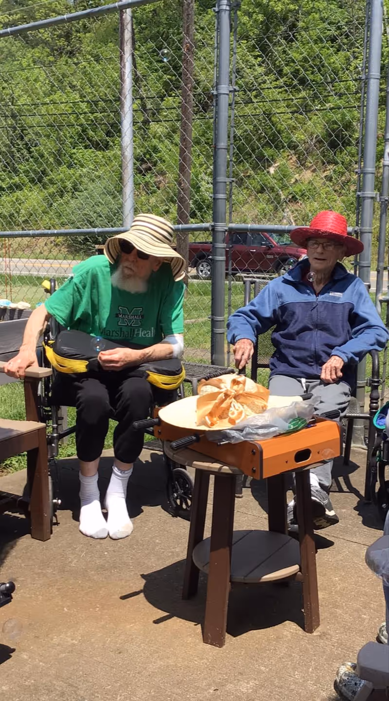 Two elderly men sitting outdoors on chairs near a chain-link fence. One man is wearing a green shirt, black pants, white socks, and a wide-brimmed striped hat, while the other man is wearing a blue jacket, gray pants, and a red hat. There is a small wooden table in front of them with a wrapped item on it. Trees and a parked vehicle are visible in the background.