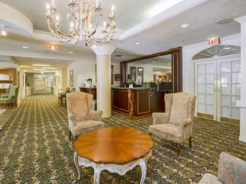 Carpeted senior living lobby with upholstered armchairs around a wooden coffee table, chandelier overhead and a reception desk beside a long hallway.